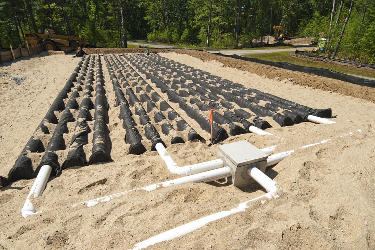 New leach field installation behind a home, showing chambers and distribution manifold with excavation equipment in background